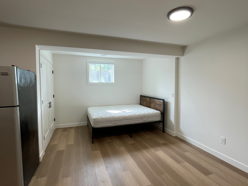 A bed sits under a small window in a studio apartment.