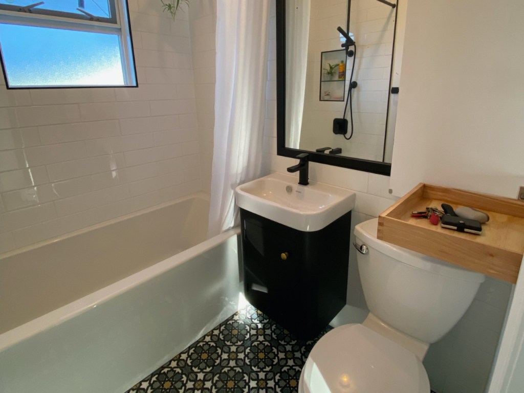 A photo of a small bathroom with a dark cabinet, dark patterned tiles, and a custom-built wooden shelf above the toilet that has been renovated by Bowden Renovations & Additions.