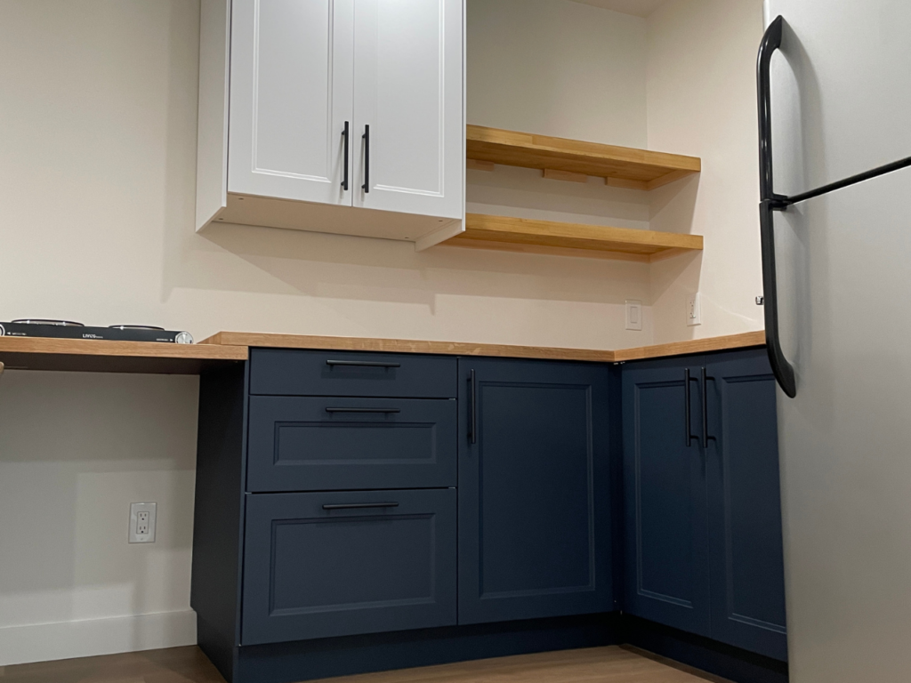 A photo of a small kitchen shows navy cupboards, light wood counters and shelves, and a refrigerator. 
