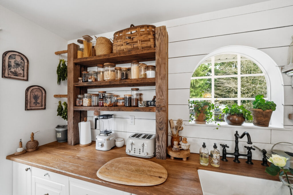 A custom-built shelving solution is built on top of the kitchen counters next to a sink that is below an arched window looking out to a backyard.
